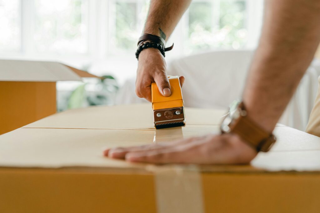 pexels-photo-4246095-4246095 Close-up of a man sealing a cardboard box with a tape dispenser during a move indoors.